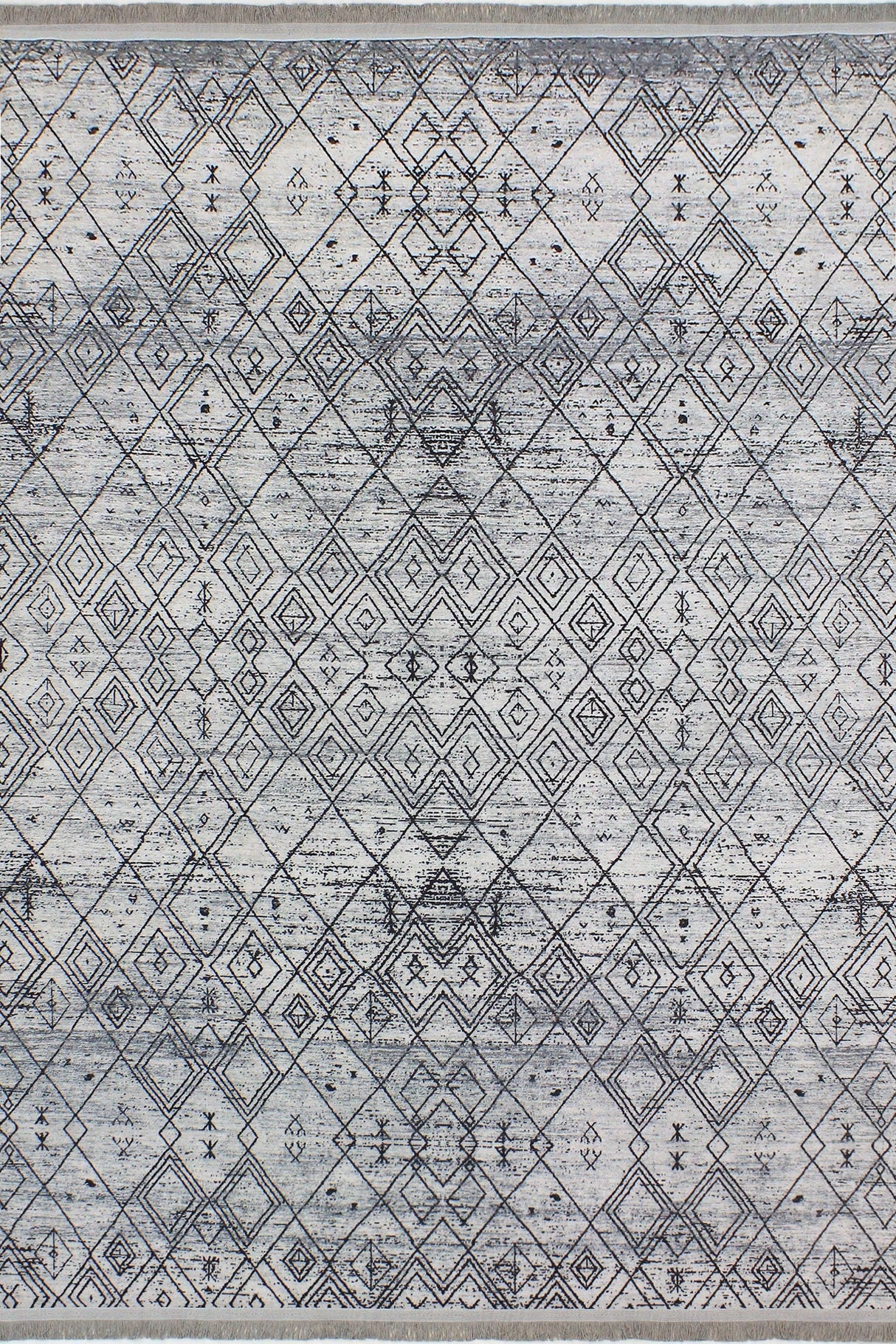 Alfombra 'Anissa' sobre fondo blanco, con un diseño geométrico en tonos grises y negros. Destacan los patrones de rombos y detalles intrincados, con un acabado sutilmente desgastado que le da un toque vintage. La alfombra es lavable en lavadora y tiene una base antideslizante.