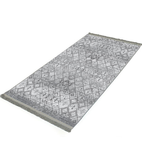 Alfombra 'Anissa' sobre fondo blanco, con un diseño geométrico en tonos grises y negros. Destacan los patrones de rombos y detalles intrincados, con un acabado sutilmente desgastado que le da un toque vintage. La alfombra es lavable en lavadora y tiene una base antideslizante.
